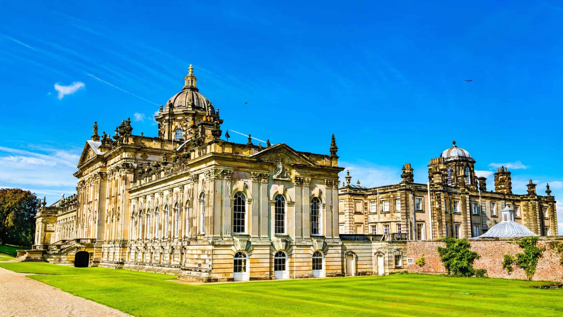 A grand historic stately home with ornate architecture under a bright blue sky and green lawns.