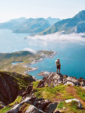 Person standing on a green cliff overlooks blue water and distant mountains under a clear sky.