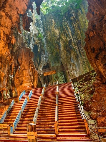 Wide stone staircase leads up inside a large, illuminated cave with a small temple and people at the base.
