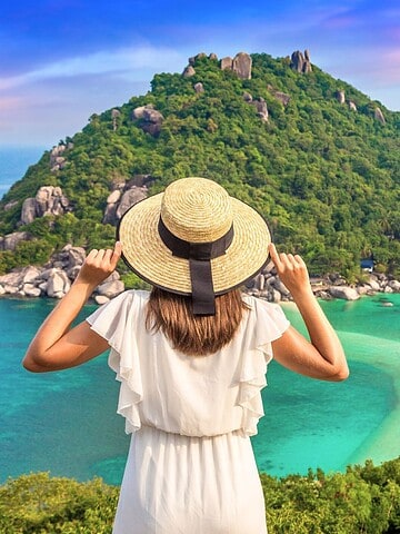 Woman in a sunhat overlooking a tropical island with turquoise water and lush greenery under a colorful sky.