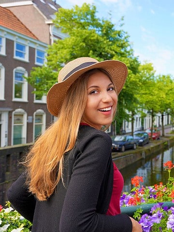 Smiling woman in a hat stands by colorful flowers on a canal bridge in a European city on a sunny day.