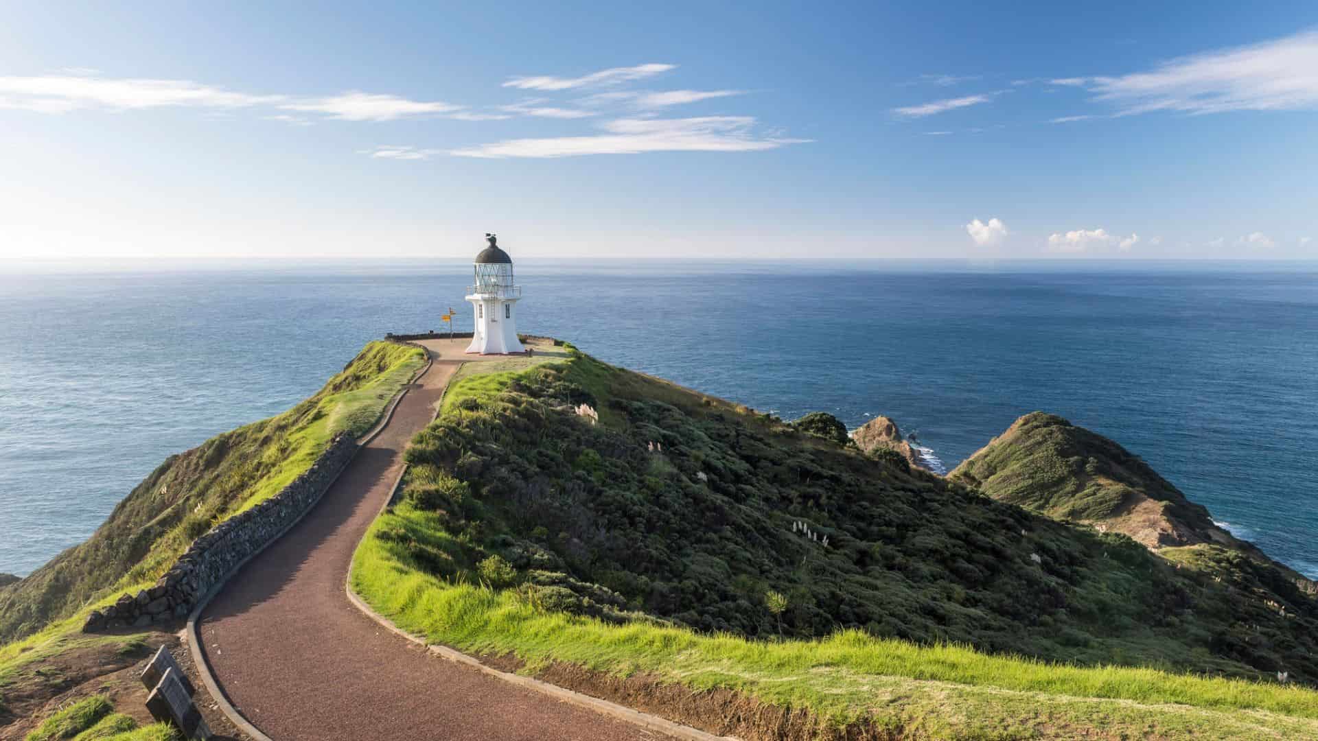 A white lighthouse stands on a grassy green hill overlooking the blue ocean under a clear sky.
