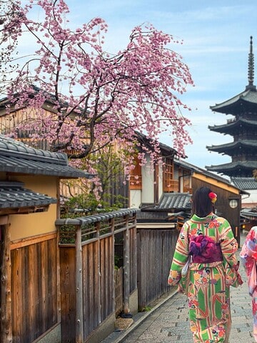 Two women in colorful kimonos walk down a traditional Japanese street with a pagoda and cherry blossoms.
