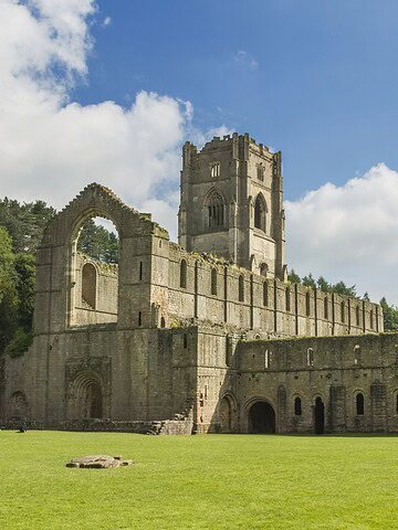 Ruins of a large stone abbey with arched windows, set on a grassy lawn under a blue sky with clouds.