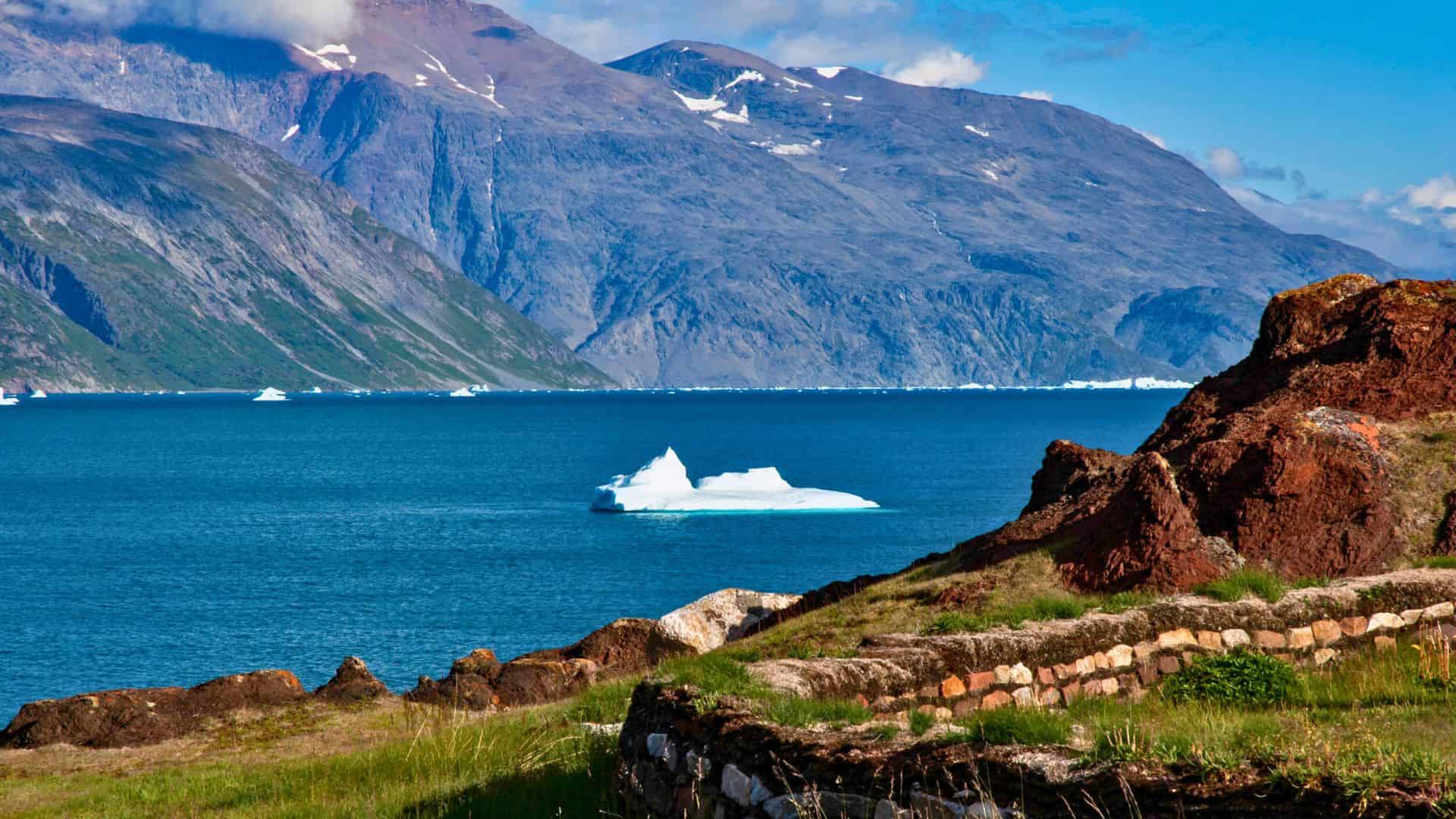 A white iceberg floats in blue water with rocky cliffs and mountains in the background.