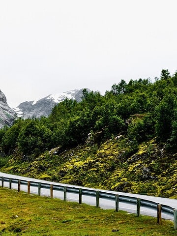Curving road with guardrail winds through green hills and mountains with patches of snow under a cloudy sky.