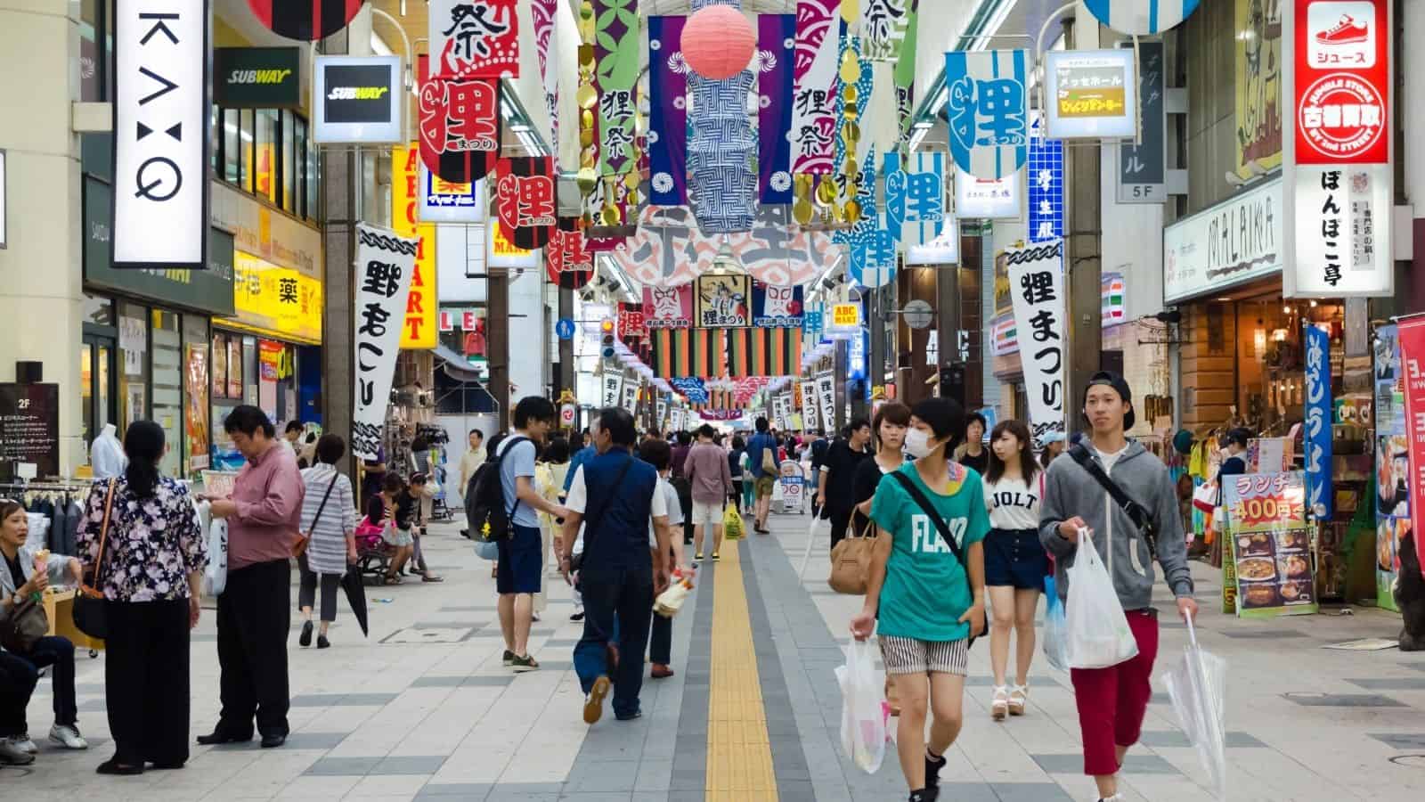 Busy Japanese shopping street with people walking and colorful banners and signs hanging overhead.