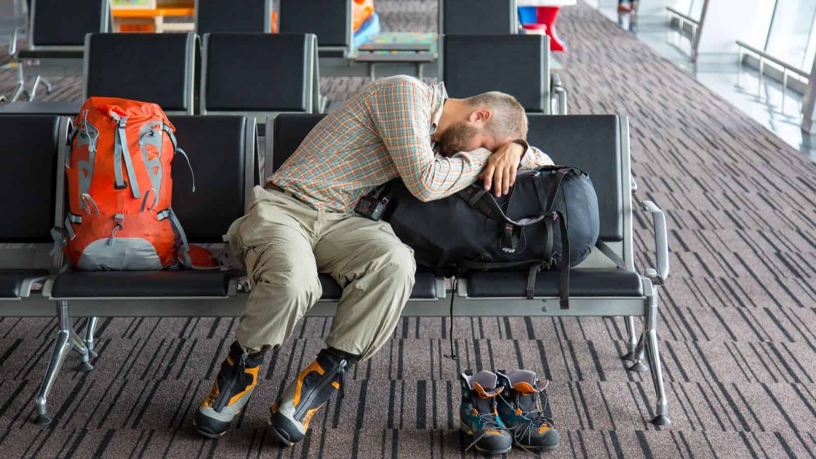 A man in hiking gear sleeps on airport seats, using his backpack as a pillow. Two backpacks and boots nearby.