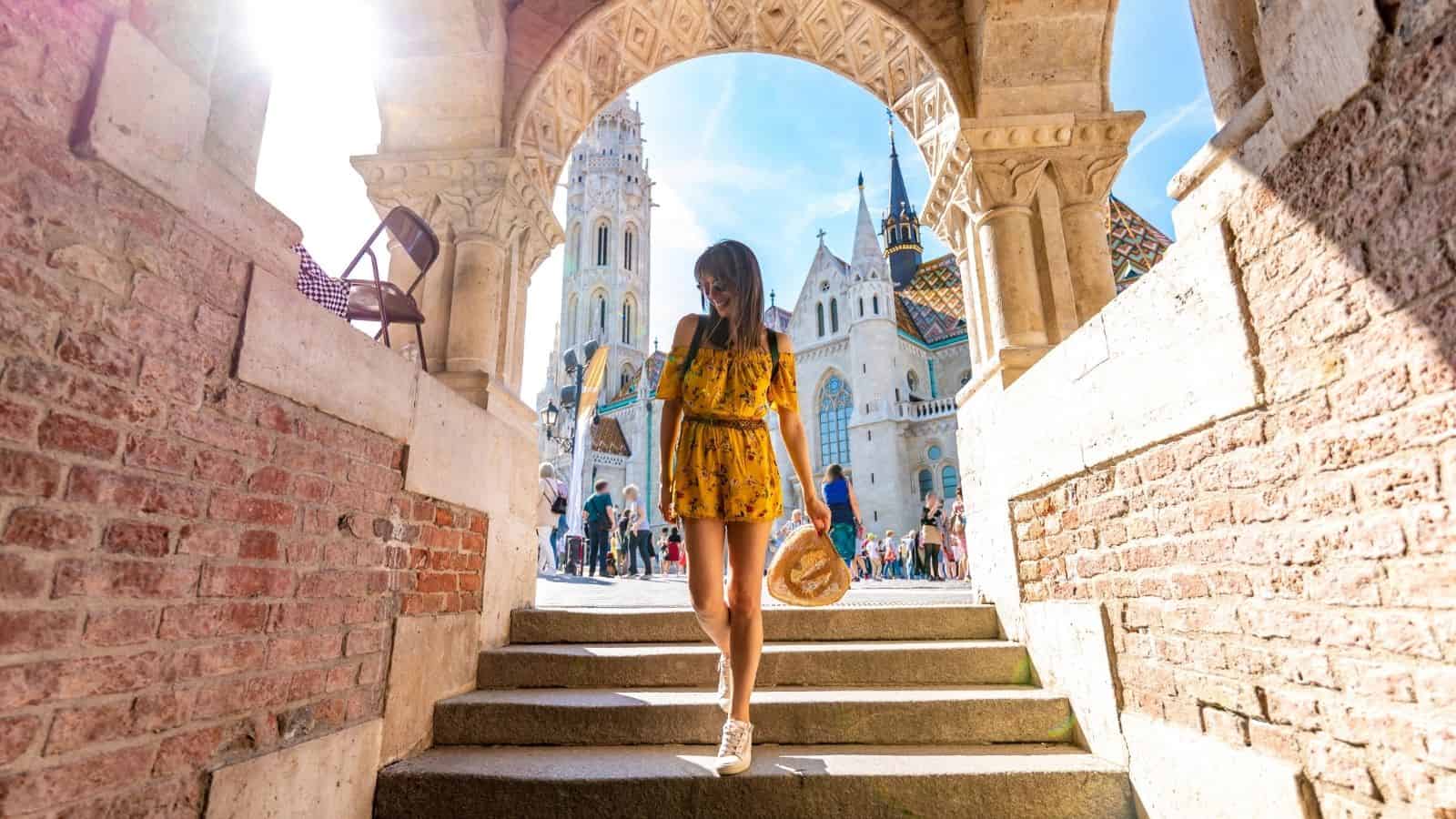 Woman in a yellow dress walks down stone steps near historic buildings under a sunny, blue sky.