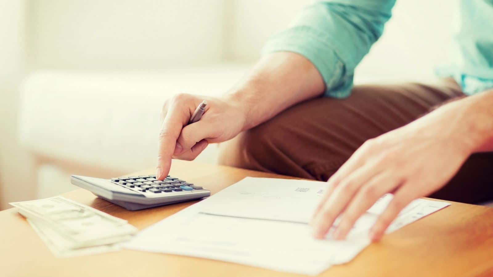 A person using a calculator with cash and papers on a table, managing finances or budgeting to avoid overspending and save for affordable trips.