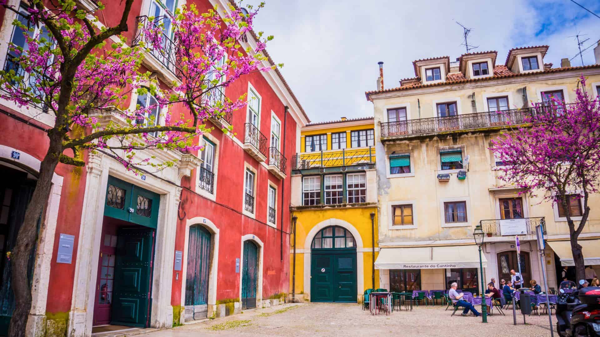 Colorful buildings and blooming trees surround a quaint outdoor caf&eacute; in a European square.