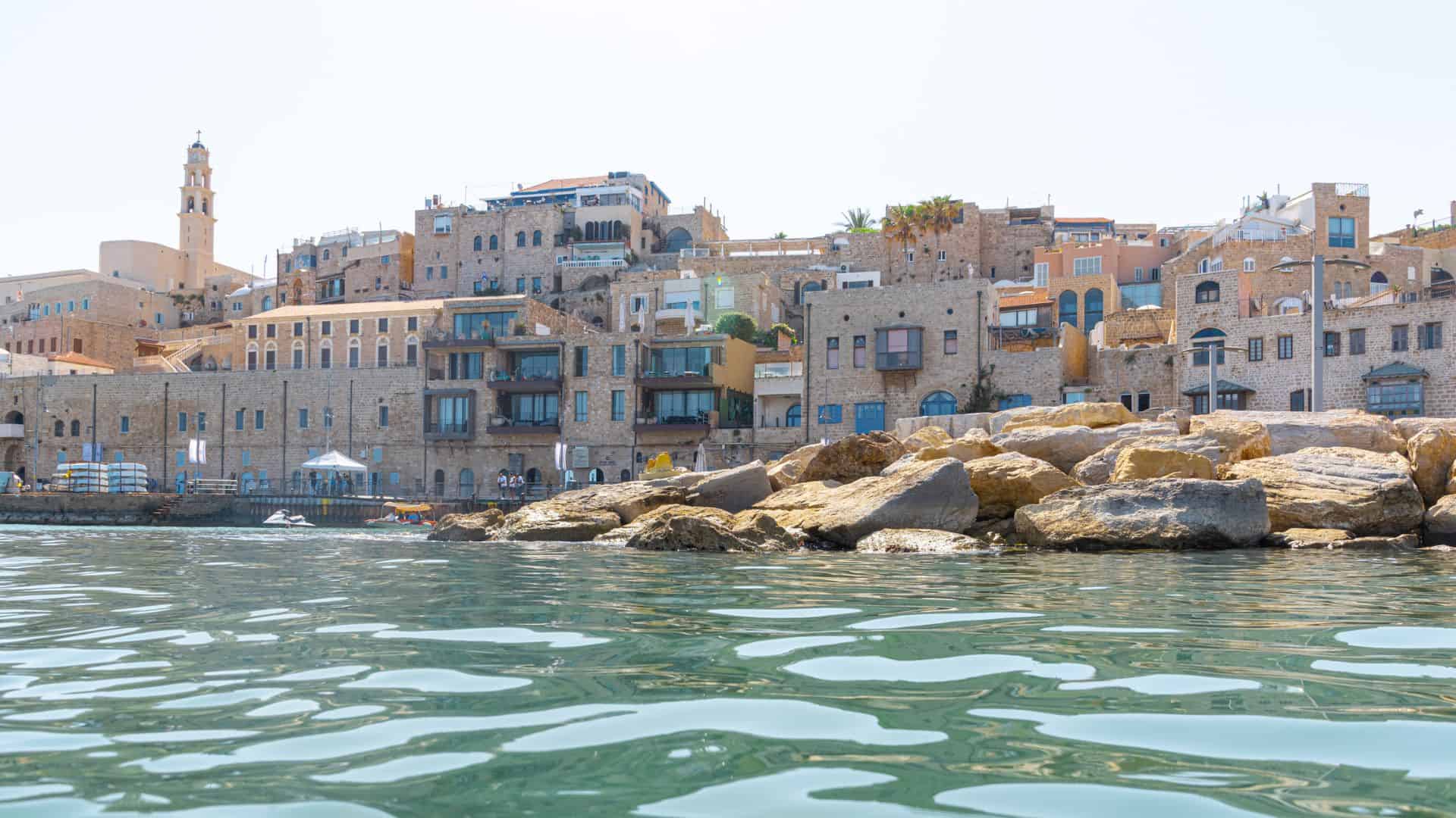 Stone buildings and houses line a waterfront with rocky shore and calm, rippling water in the foreground.