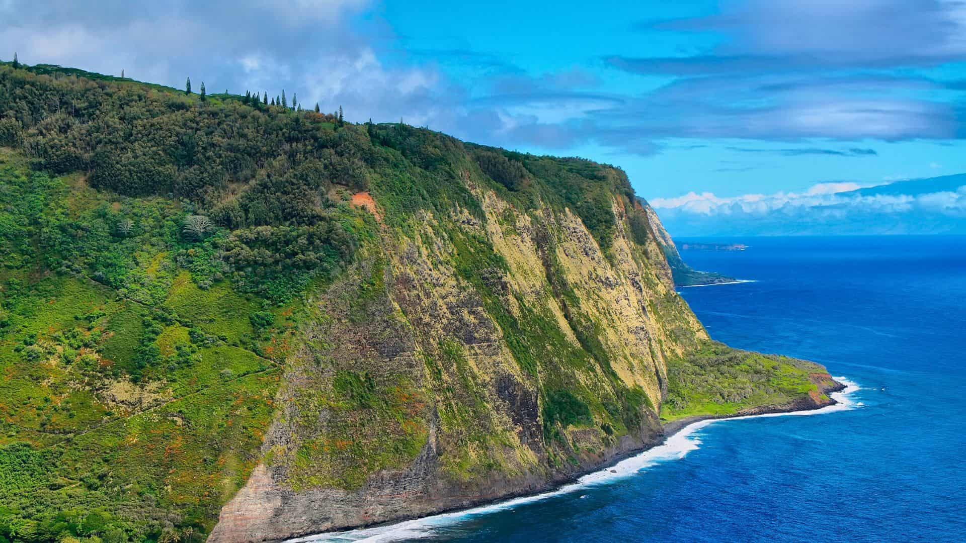 Steep green cliffs overlook a deep blue ocean under a partly cloudy sky.