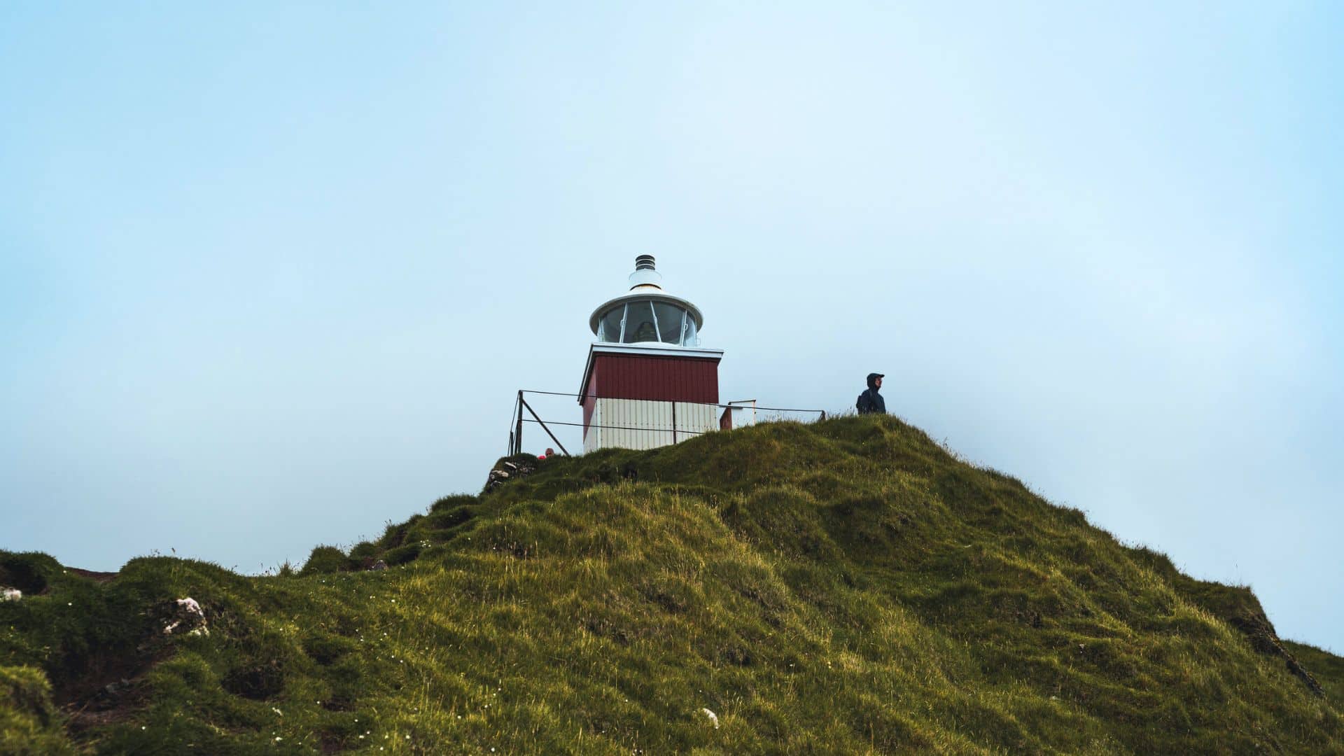 A small lighthouse sits atop a grassy hill with a person standing nearby under a cloudy sky.