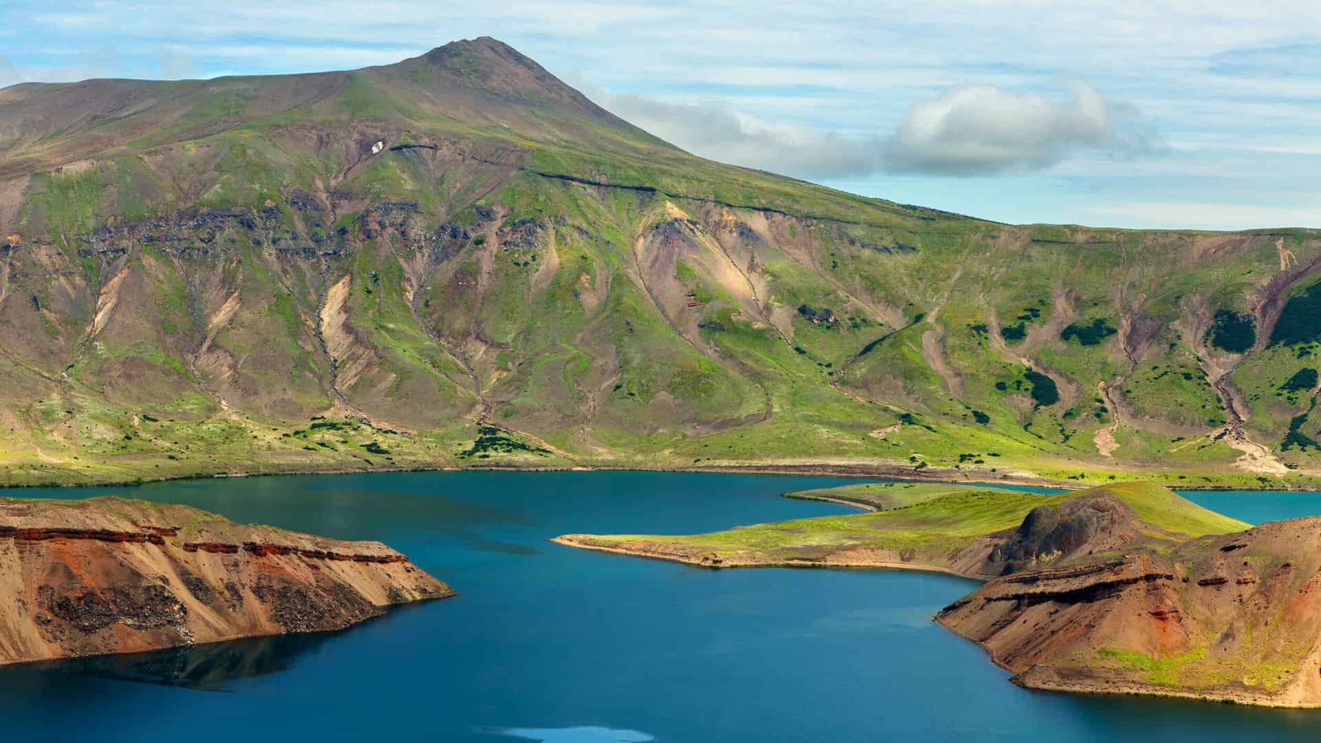 A blue lake surrounded by green hills and a mossy mountain under a partly cloudy sky.