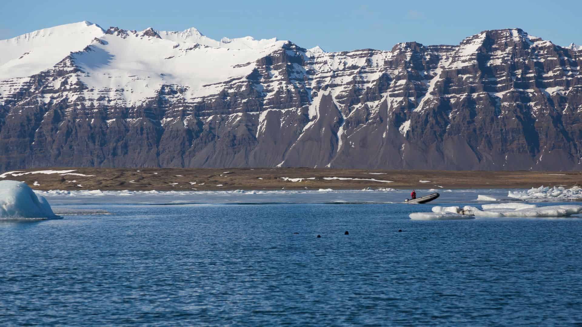 A small boat on blue water with icebergs, and snow-capped mountains in the background.