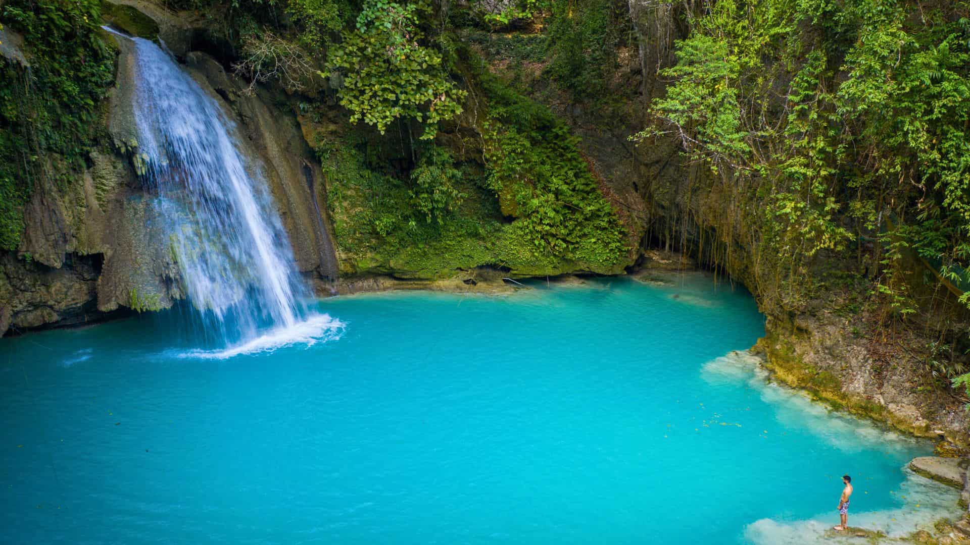 A turquoise waterfall pool surrounded by lush greenery; a person stands at the water&rsquo;s edge.