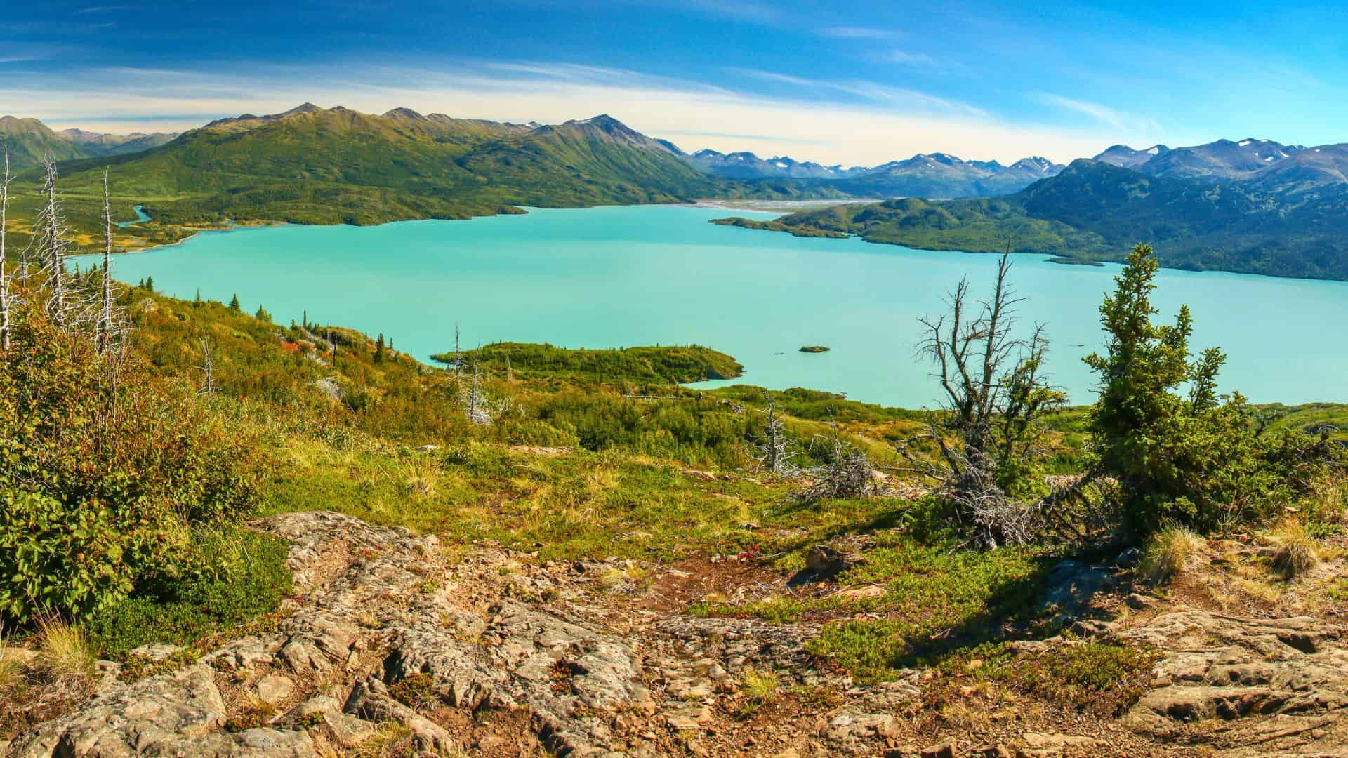 A turquoise lake surrounded by green hills and distant mountains under a clear blue sky.