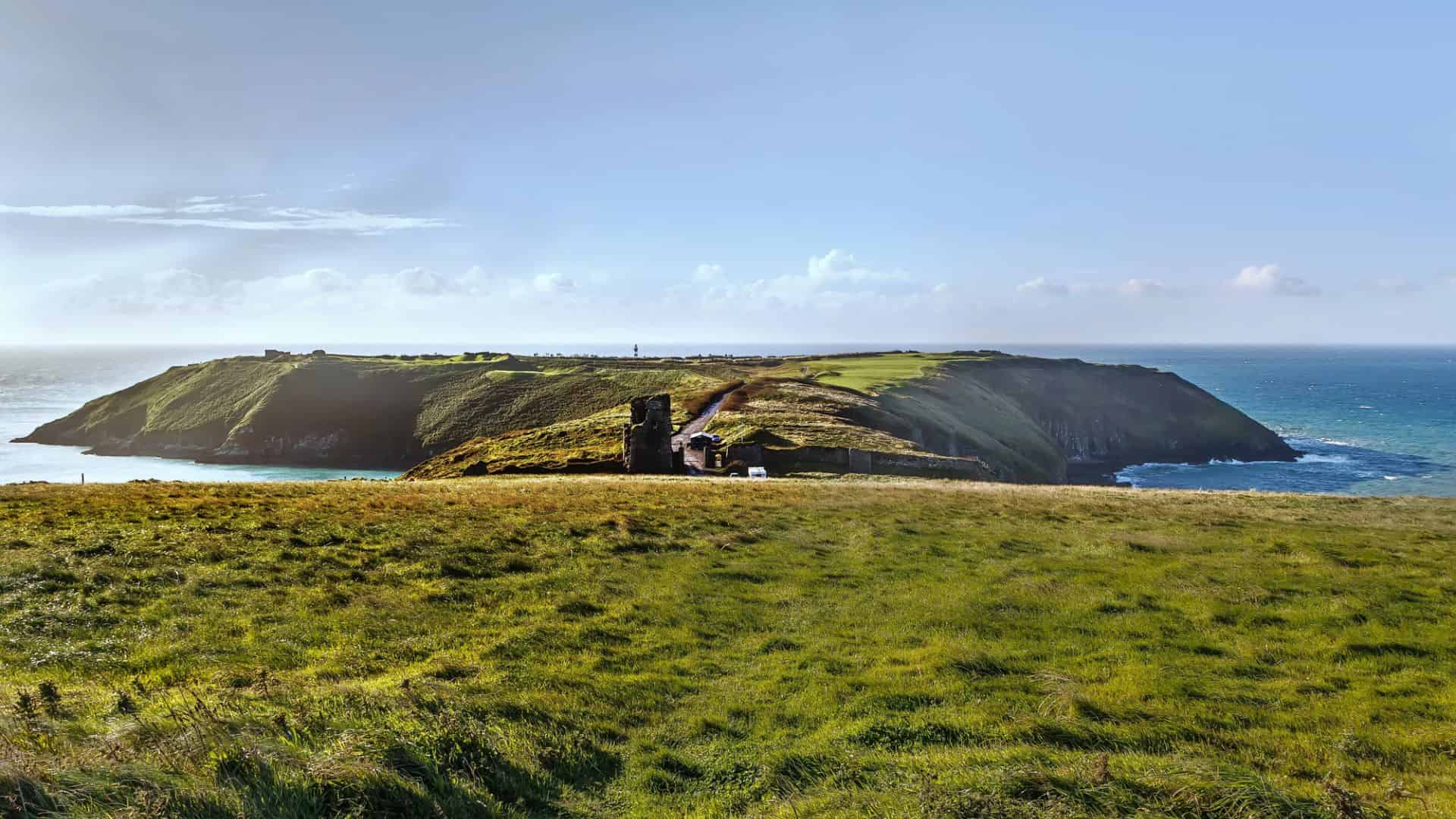 Grassy cliffs overlooking the sea with a road and ruins, under a blue sky with scattered clouds.