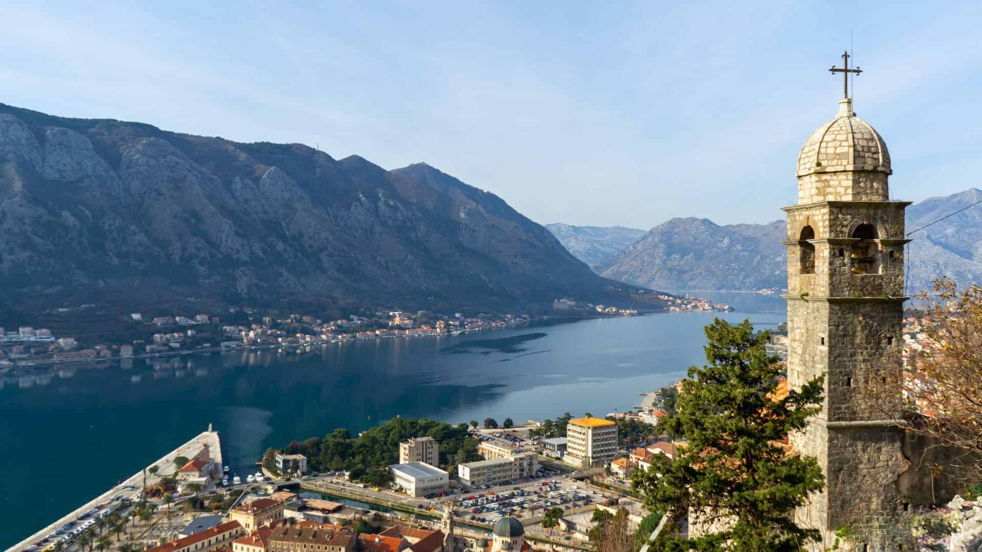 A stone bell tower overlooks a bay surrounded by mountains and a coastal town under a clear sky.