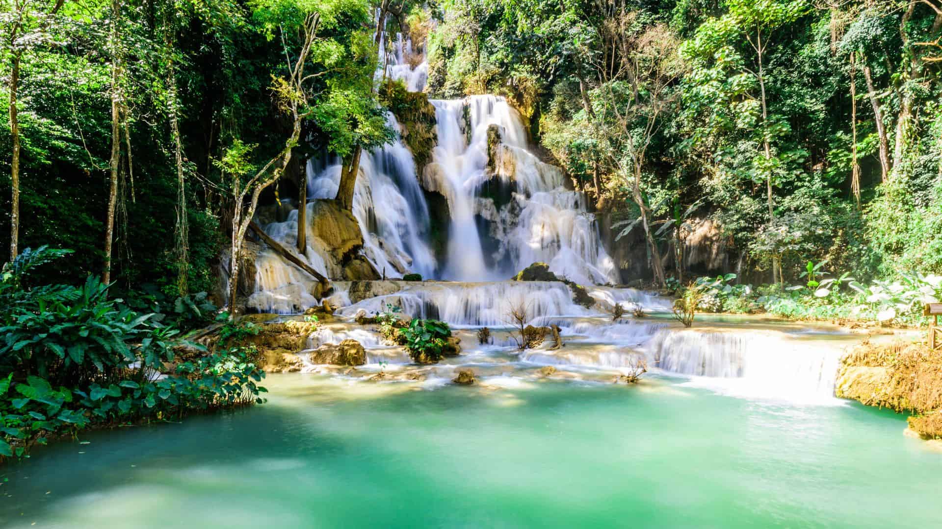 A multi-tiered waterfall cascades into a turquoise pool surrounded by lush green forest.