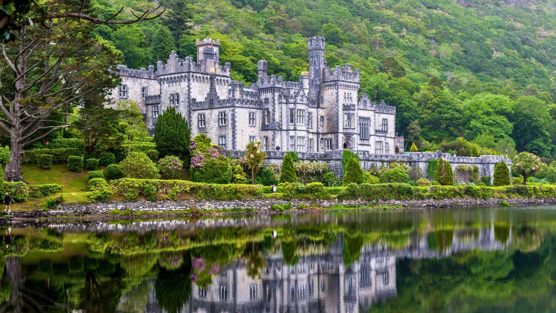 A grand stone castle surrounded by greenery is reflected in a calm lake in front of it.