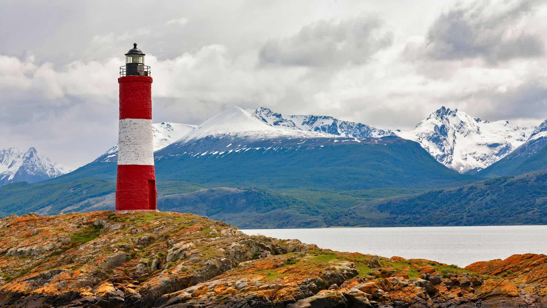 Red and white lighthouse on rocky shore with snowy mountains and water in the background under cloudy skies.