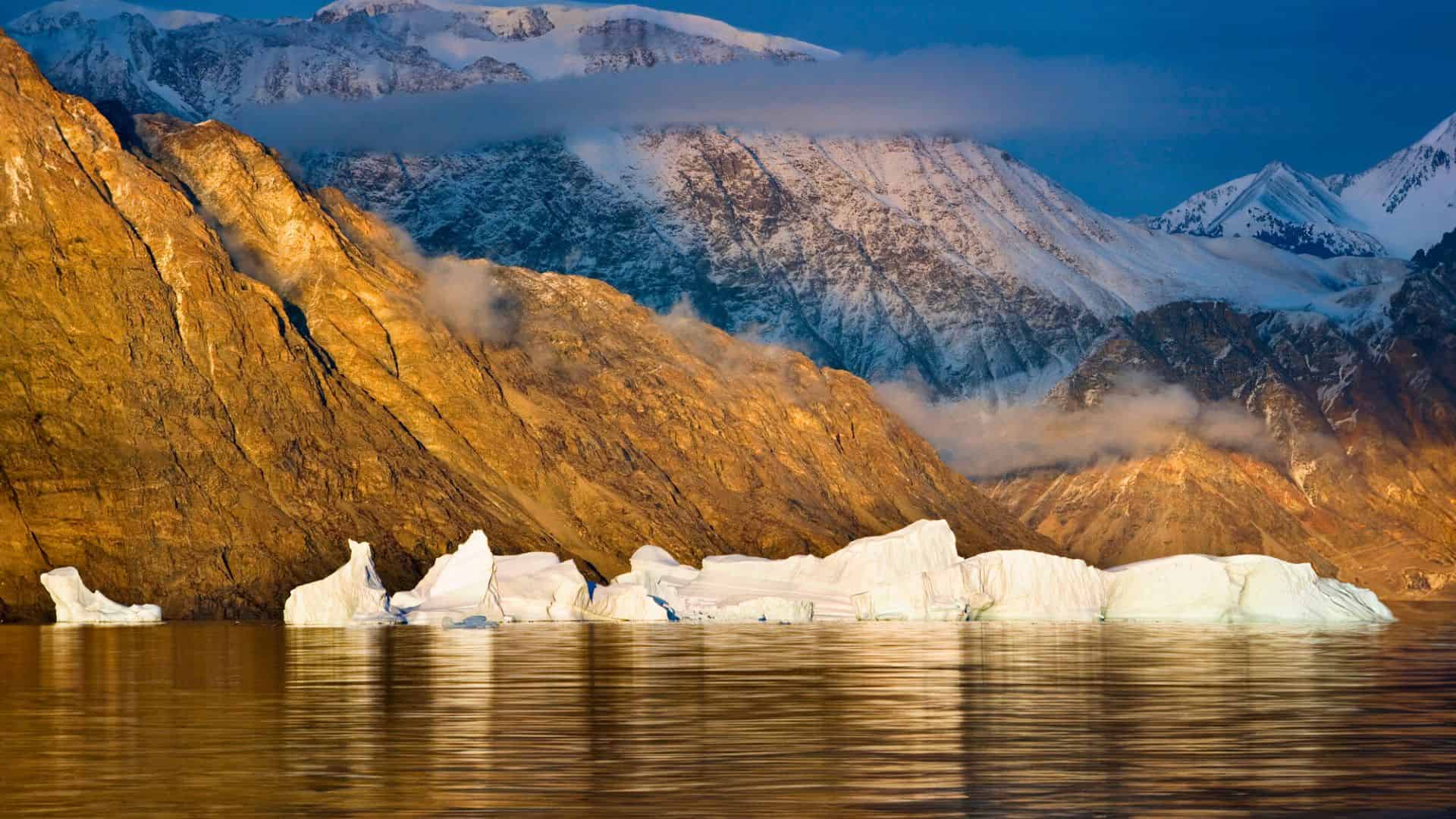 Snowy mountains and icebergs reflected in calm water under golden sunlight and clouds.