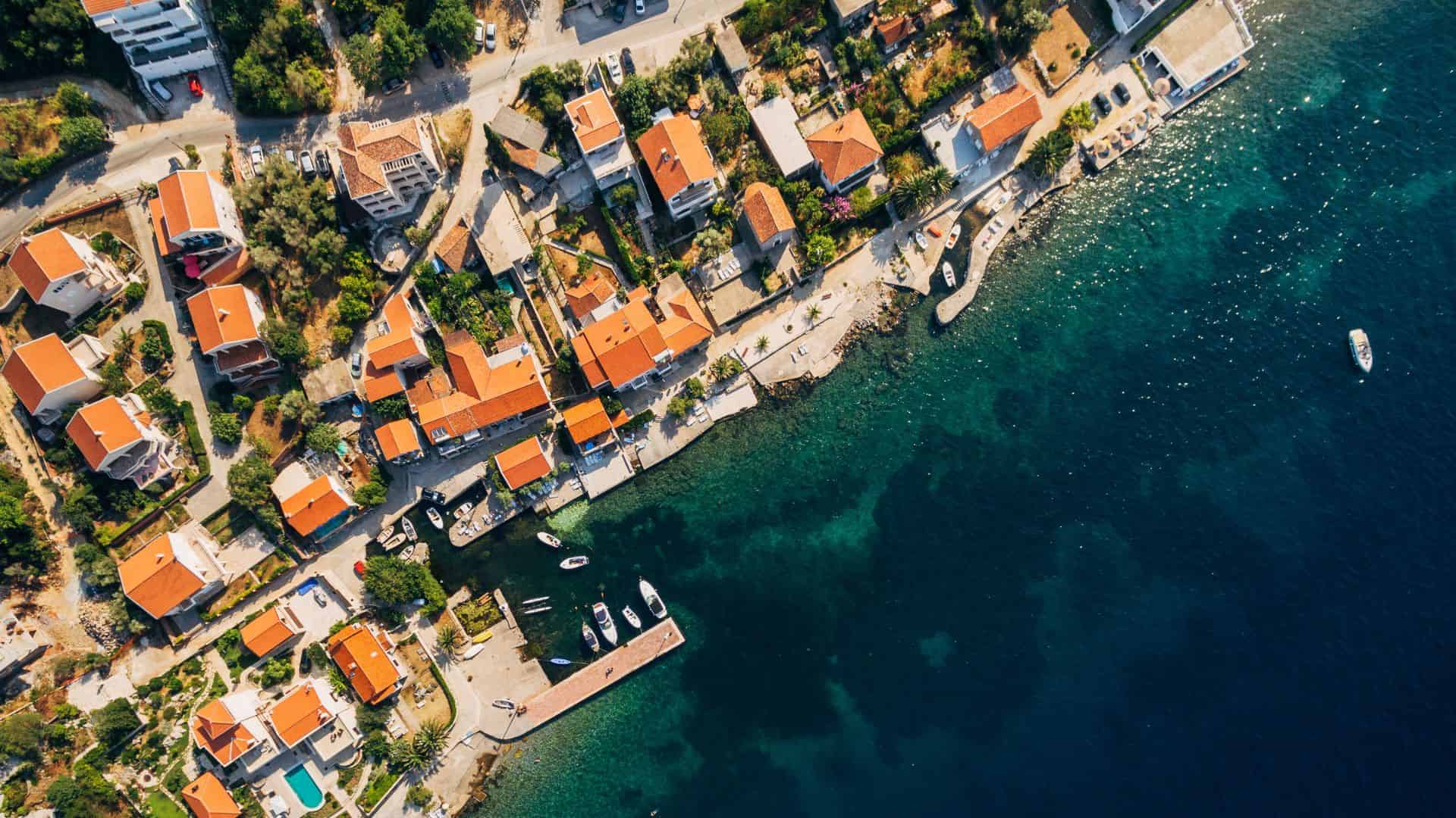 Aerial view of coastal village with red-roofed houses, a dock, and boats along clear blue water.