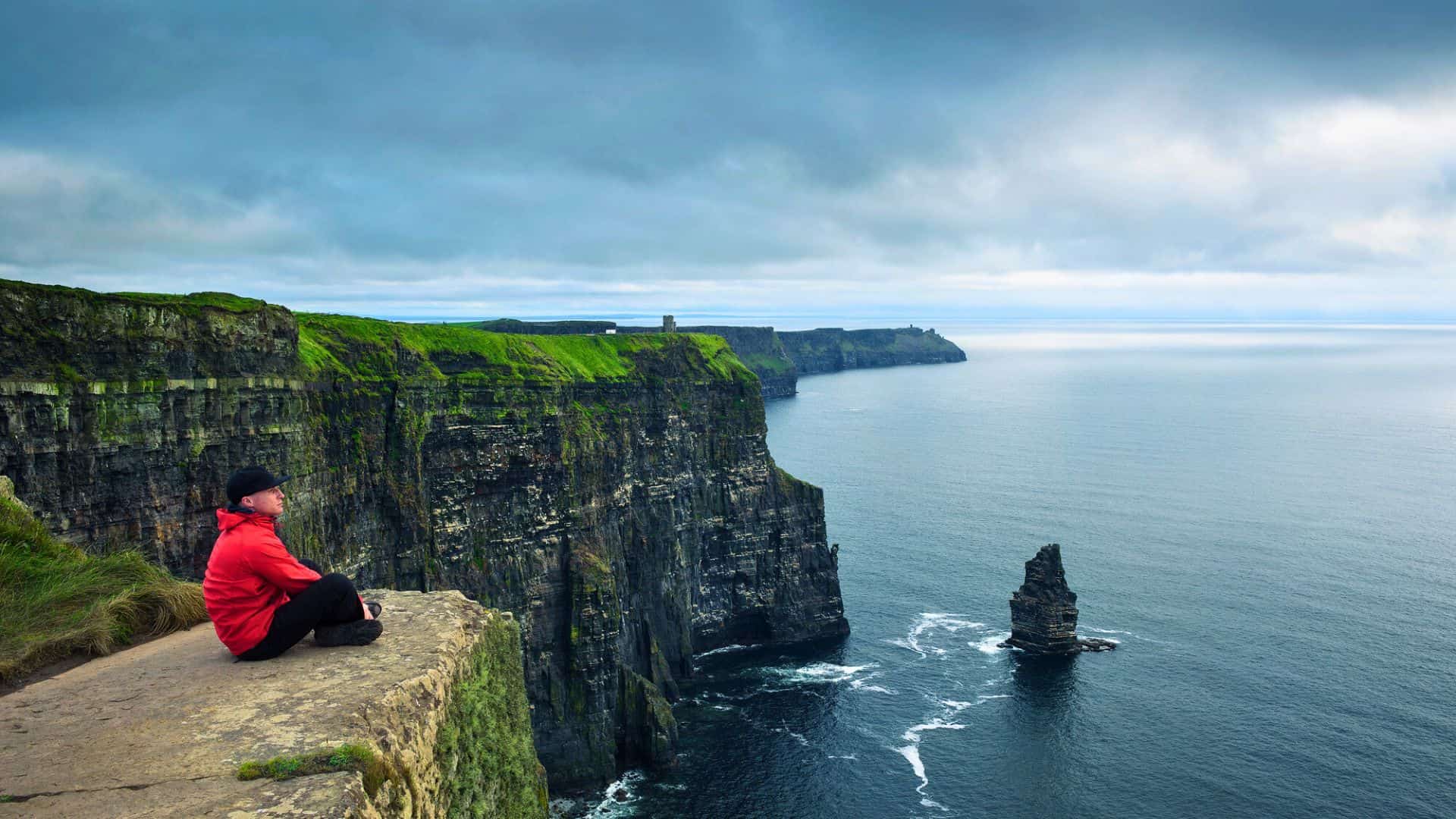 Person in a red jacket sits on a cliff edge overlooking the ocean and the Cliffs of Moher in Ireland.
