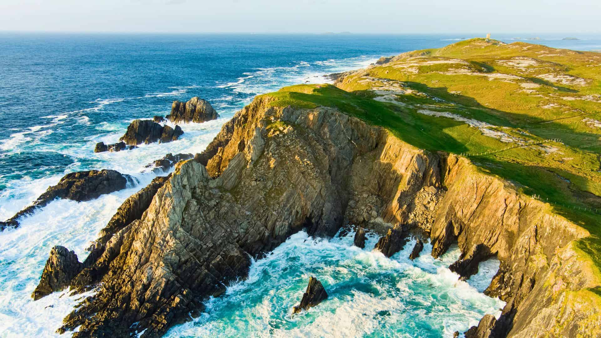 Aerial view of rugged cliffs and waves crashing along a green, rocky coastline under a clear sky.
