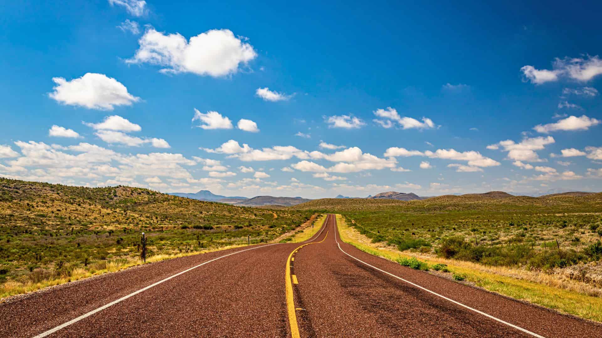 Straight empty road stretches through green landscape under a bright blue sky with scattered clouds.