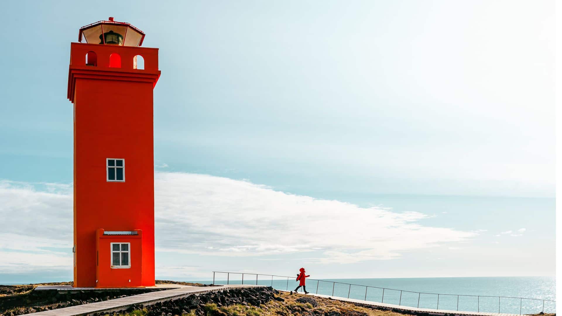Bright red lighthouse near the ocean, with a person walking on a path under a clear, sunny sky.