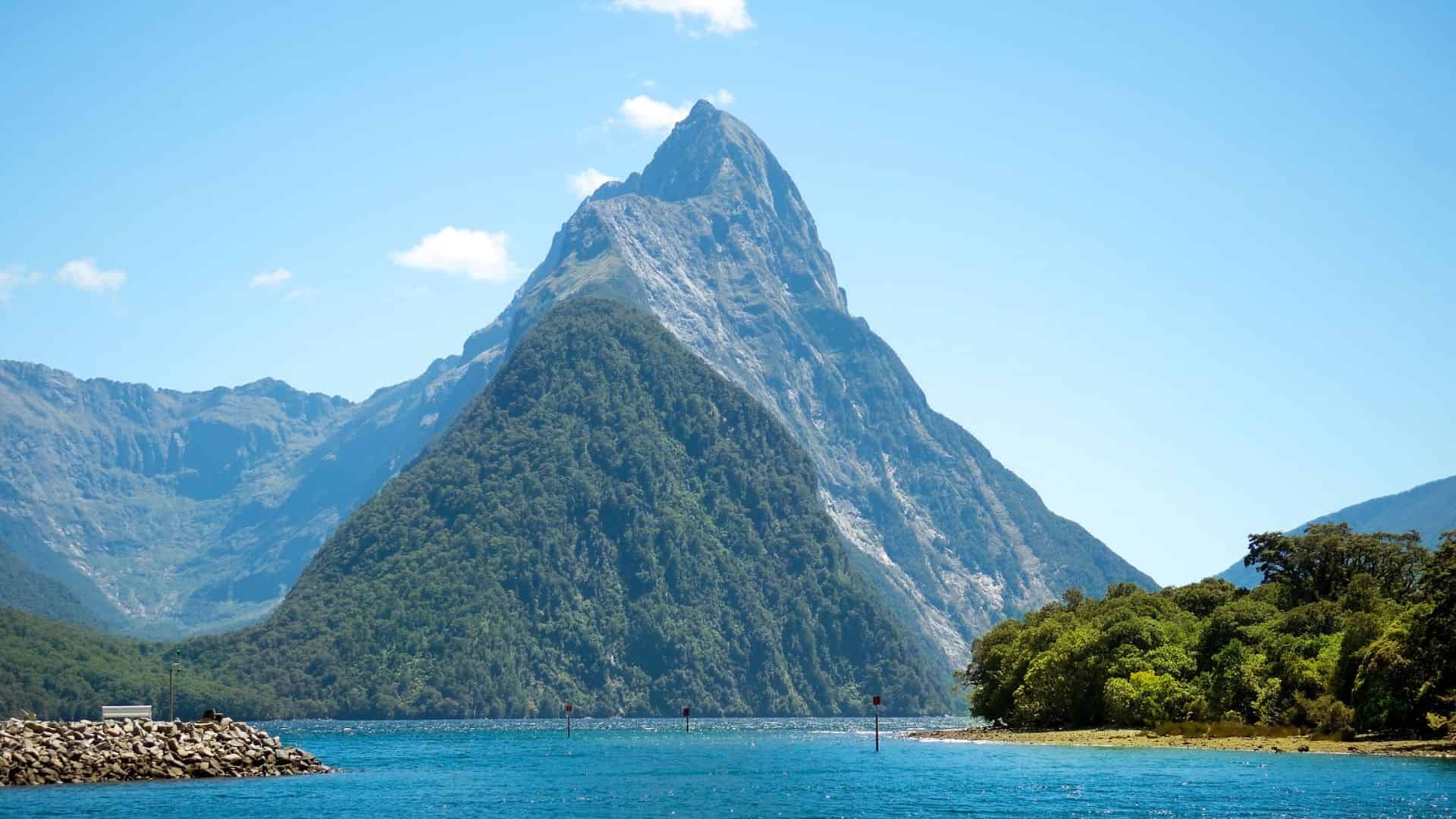A tall, green mountain rises above a blue lake under a clear, sunny sky.