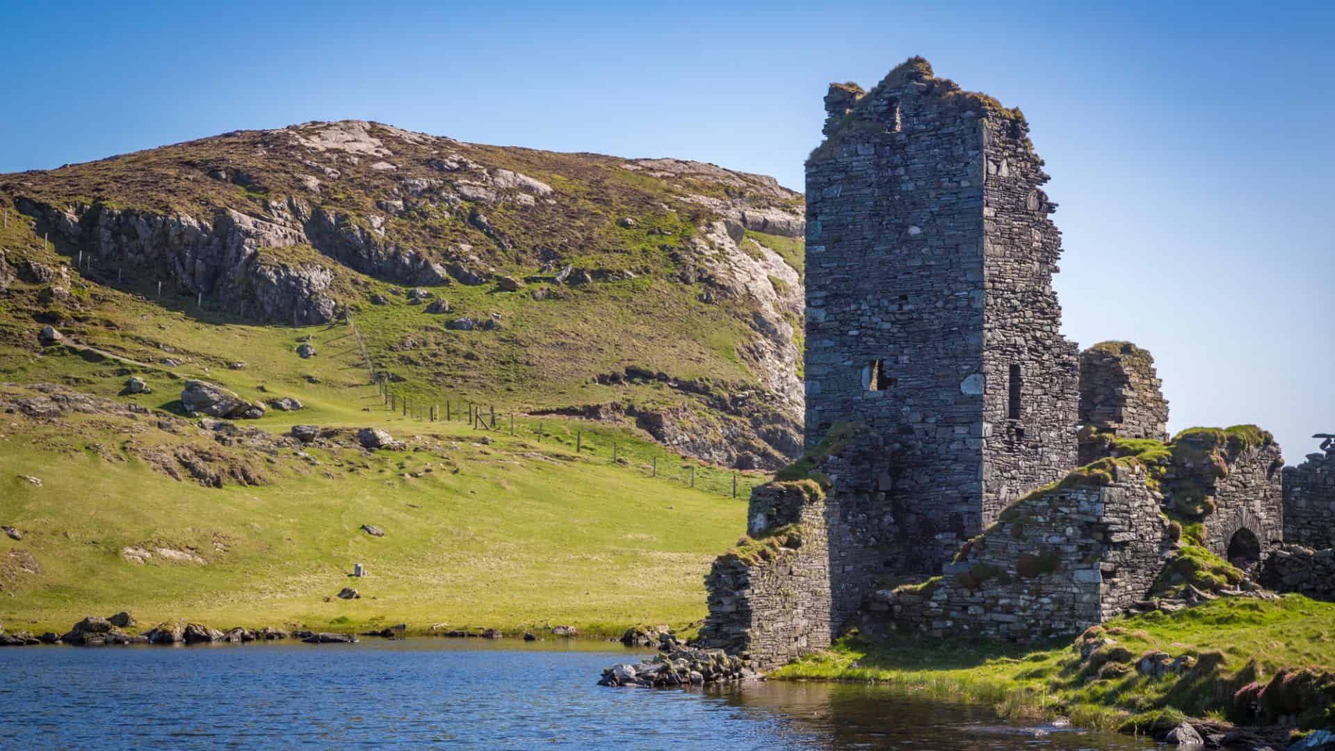 Ruins of an old stone tower by a lake, with hills and grassy landscape in the background under a blue sky.