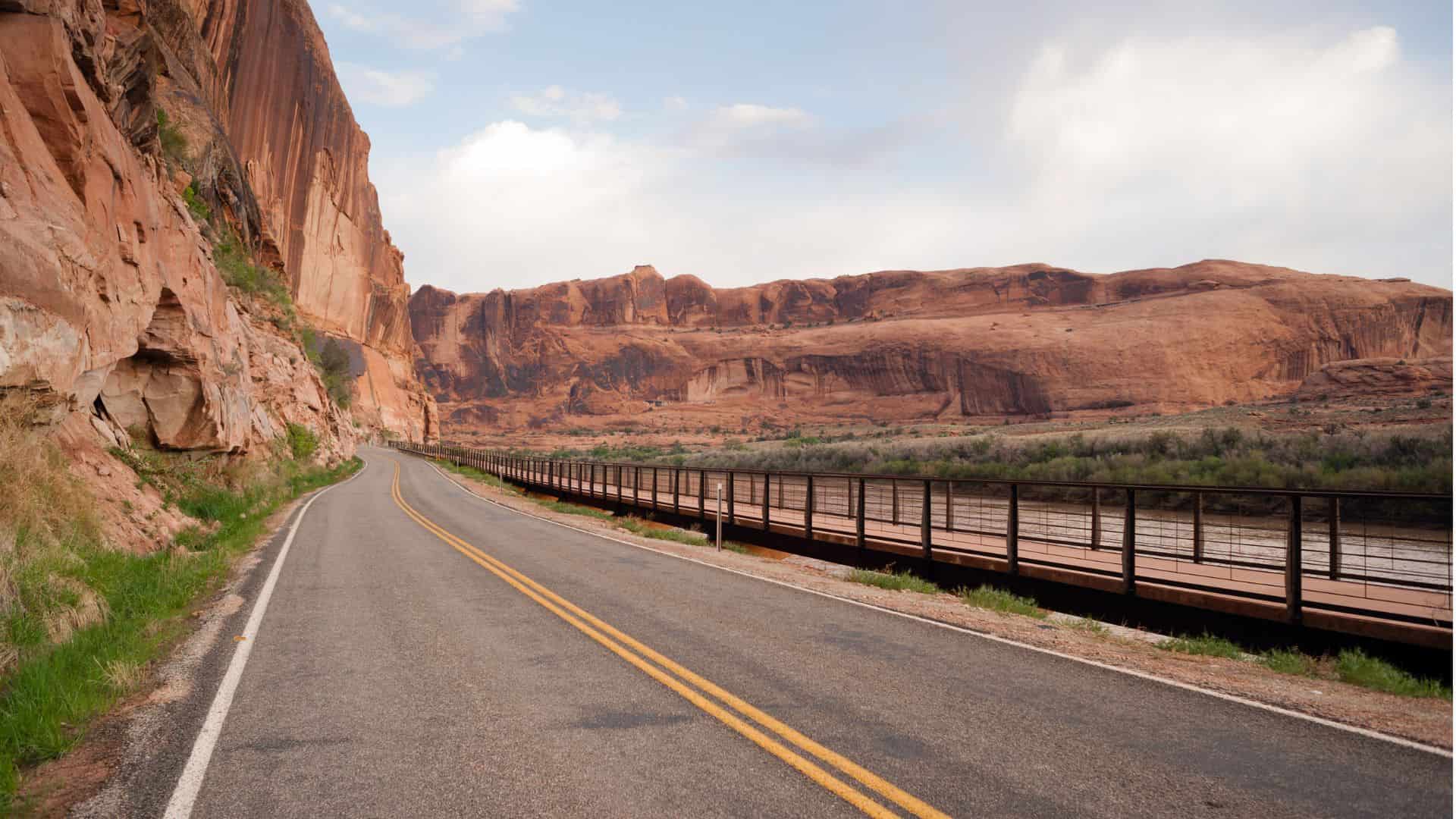 Desert road curves past red rock cliffs and a river, with a metal guardrail and cloudy sky overhead.