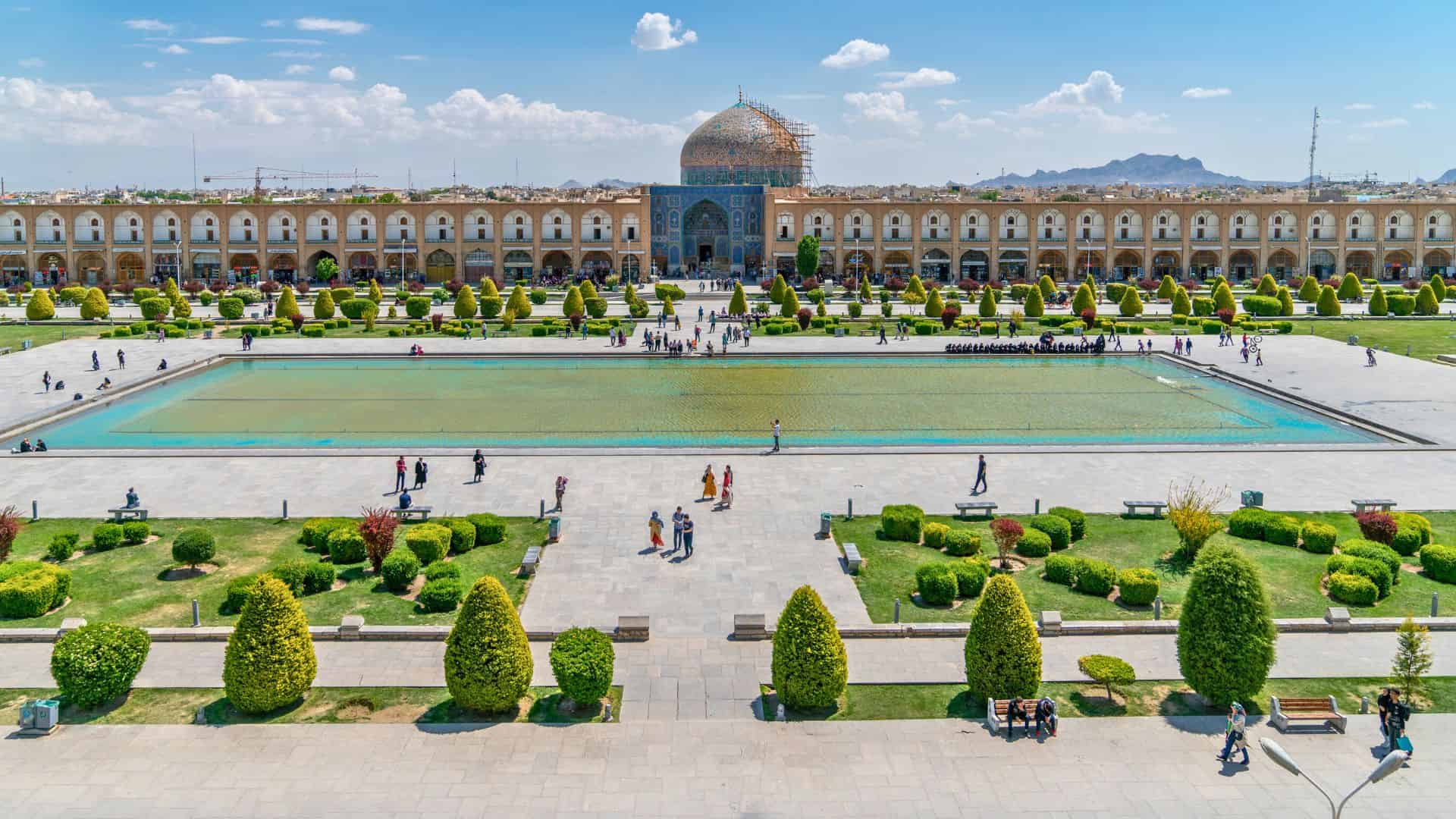 A large mosque with a blue dome faces a long reflecting pool and manicured gardens filled with people.