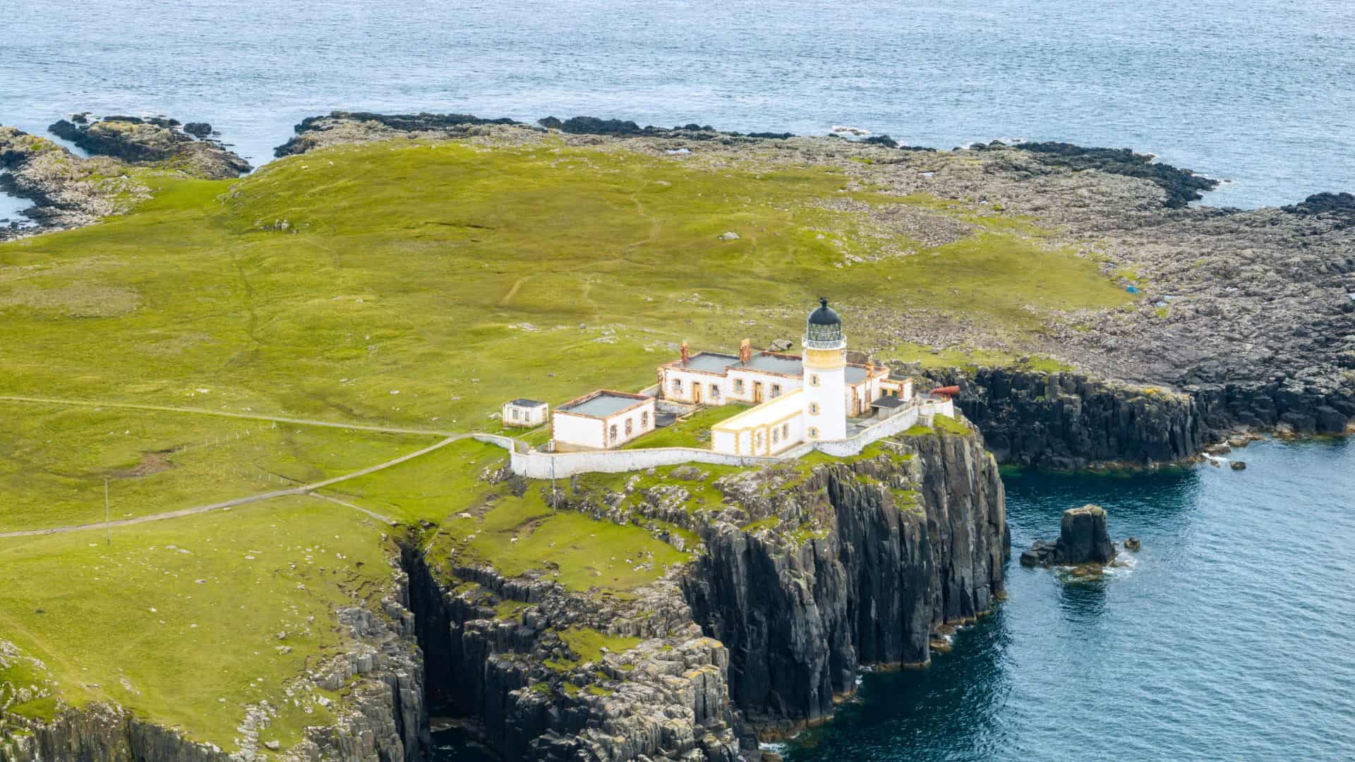 A lighthouse and buildings sit on a grassy cliff overlooking the ocean, surrounded by rocky terrain.
