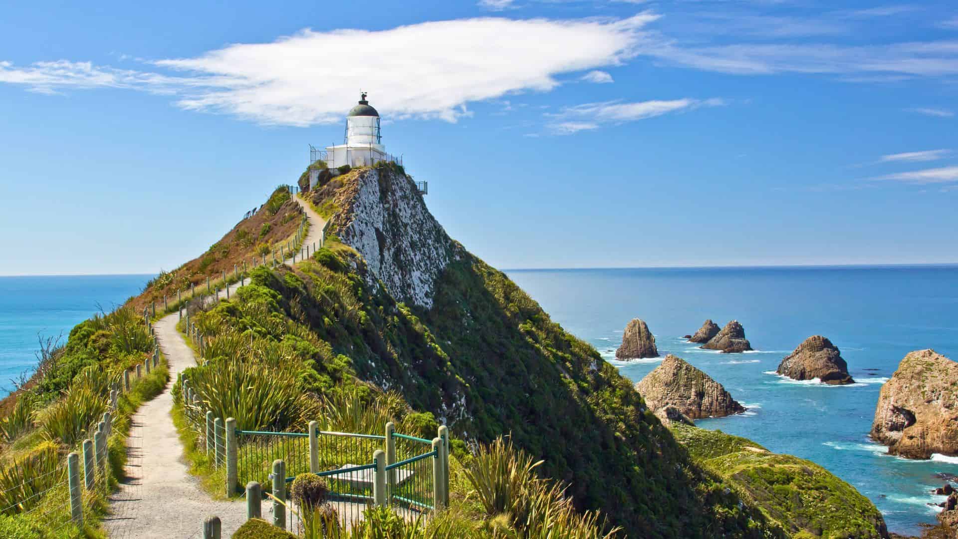 A lighthouse sits atop a green hill overlooking rocky islets and the blue ocean under a sunny sky.