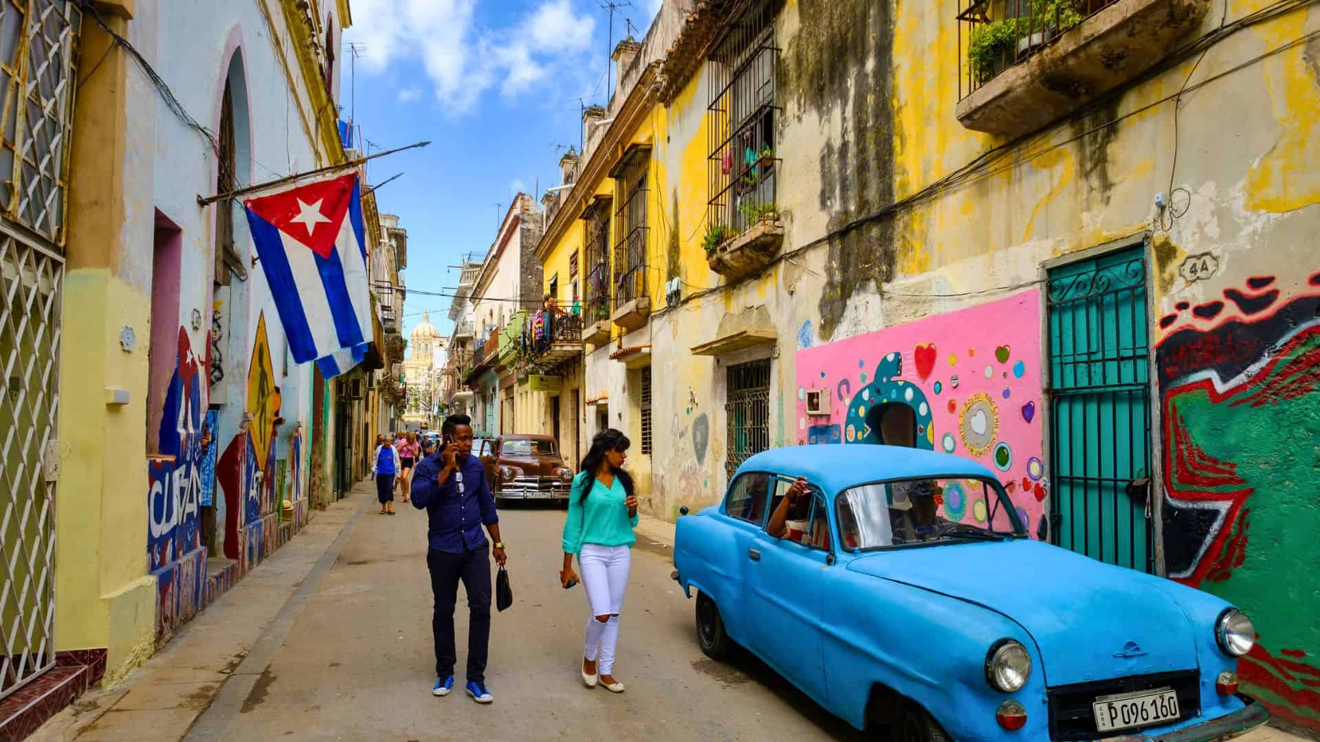 People walk past colorful murals, a blue vintage car, and a Cuban flag on a vibrant street in Havana.