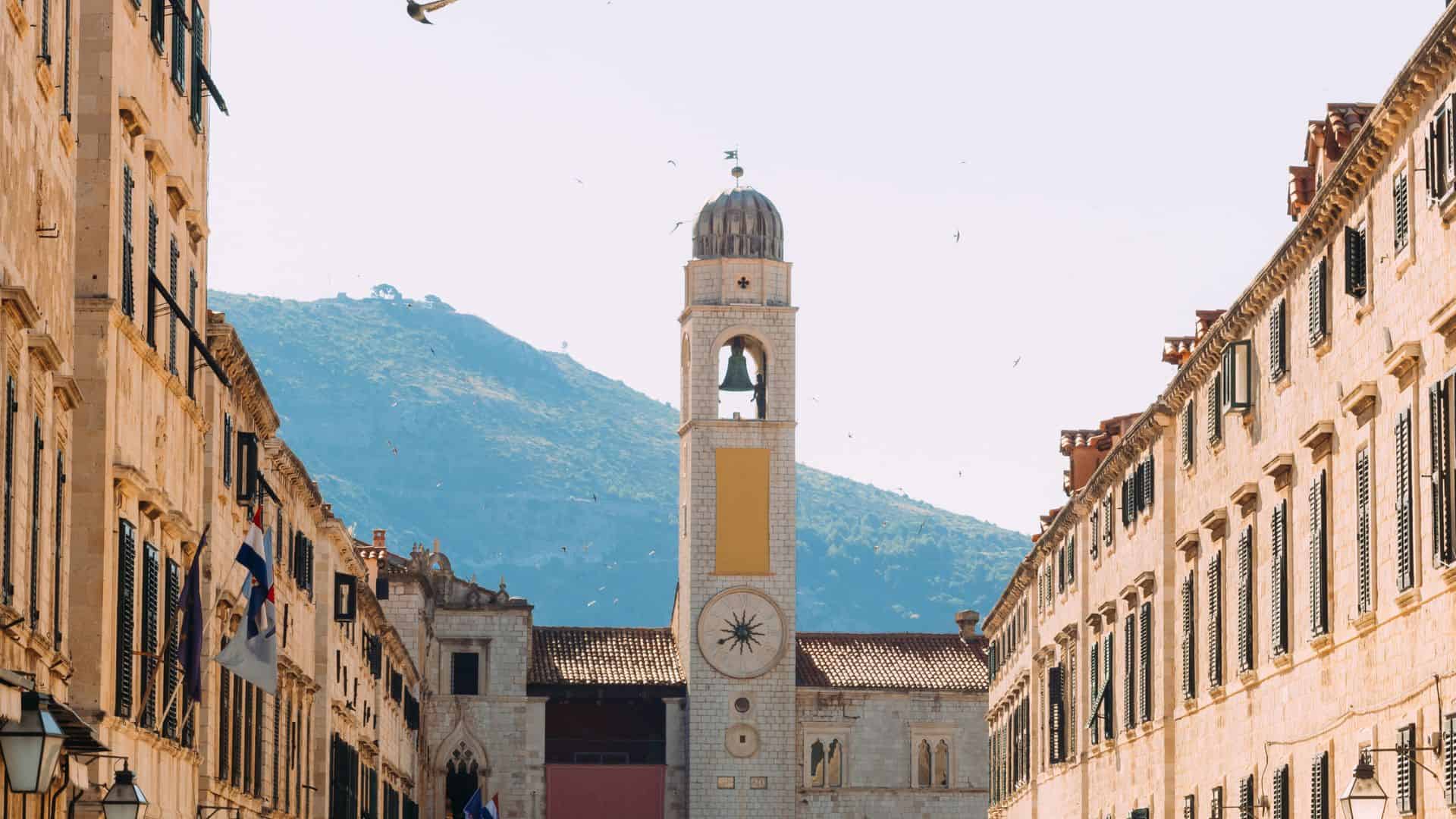 A bell tower stands between rows of old buildings with mountains in the background on a sunny day.