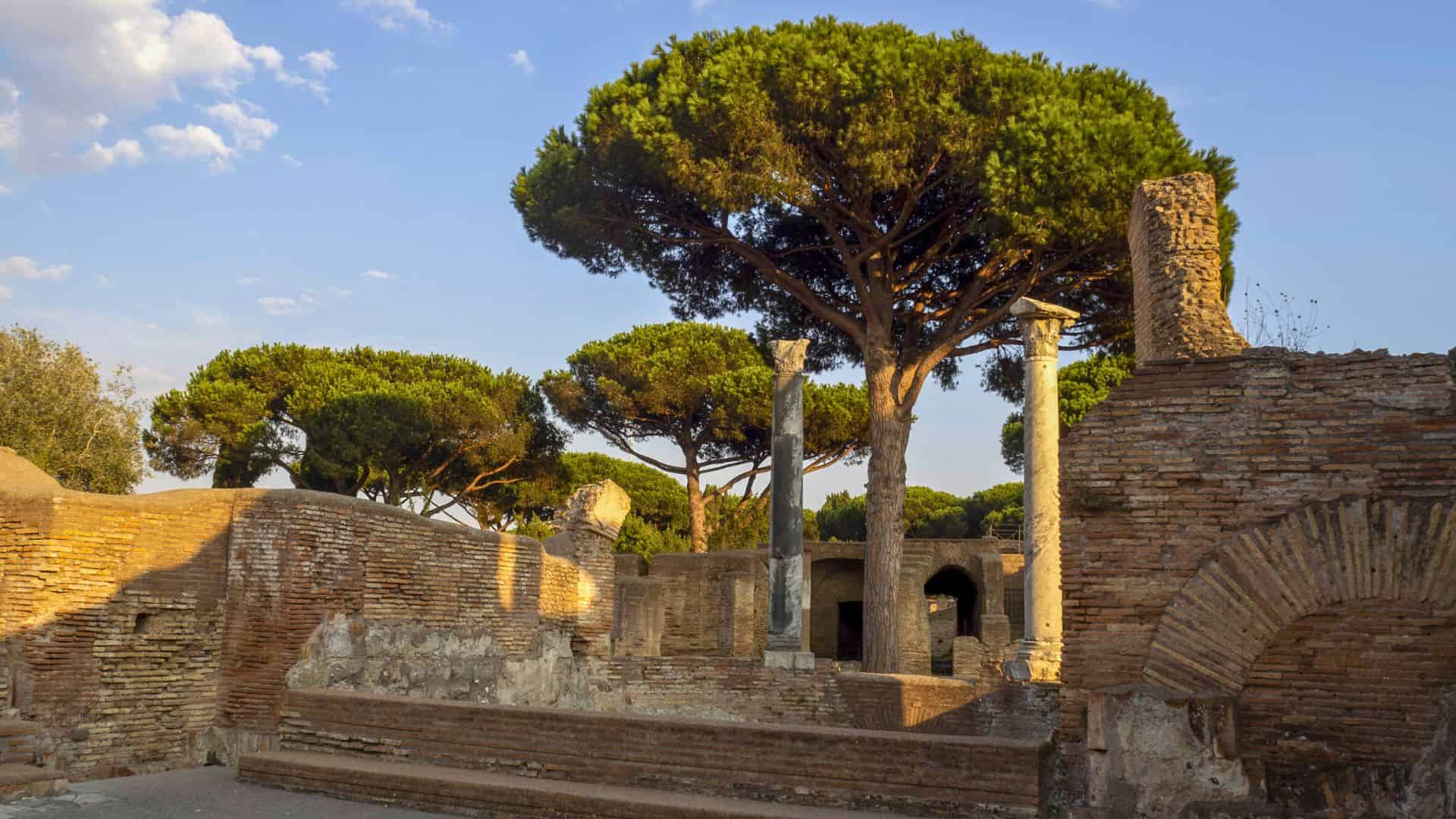 Ancient brick ruins with stone columns and large pine trees under a blue sky with scattered clouds.