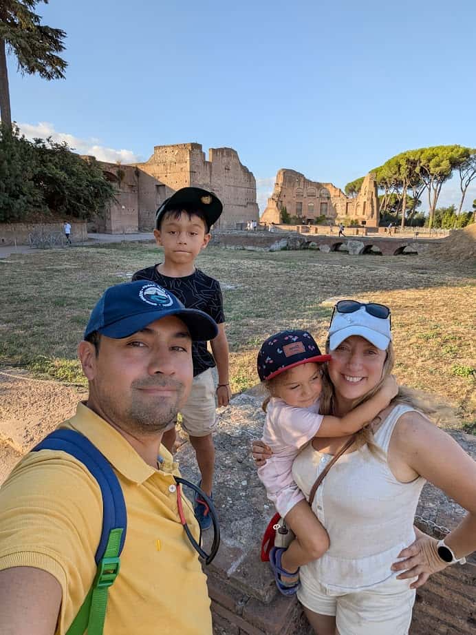 Family of four smiling and posing for a selfie at ancient ruins on a sunny day, creating memories in Rome with kids.