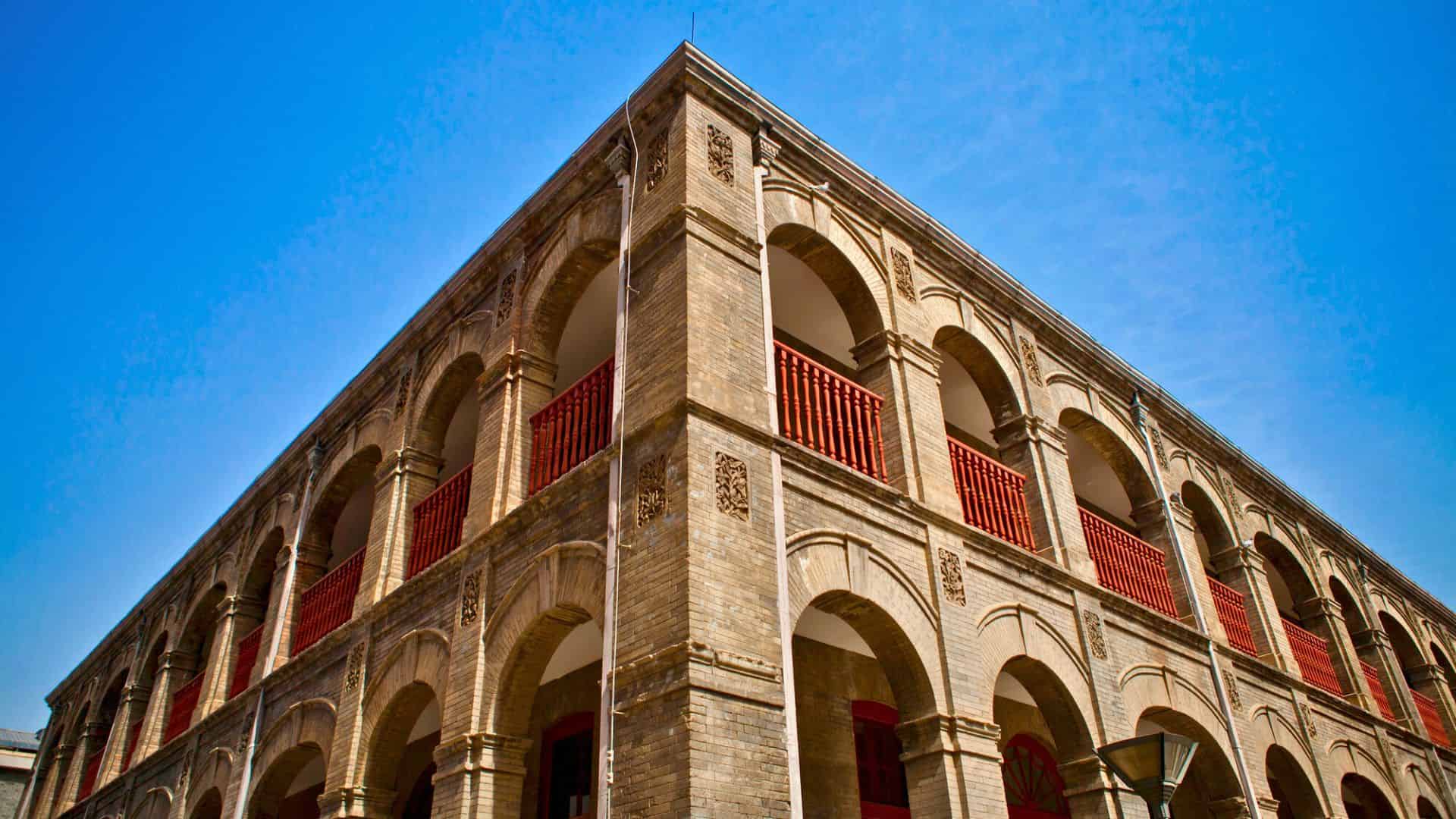 A historic brick building with arches and red railings against a bright blue sky.