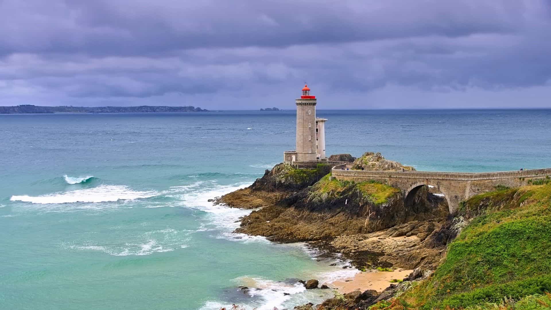 A lighthouse on a rocky coast under a cloudy sky, with waves crashing and greenery on the cliffs.