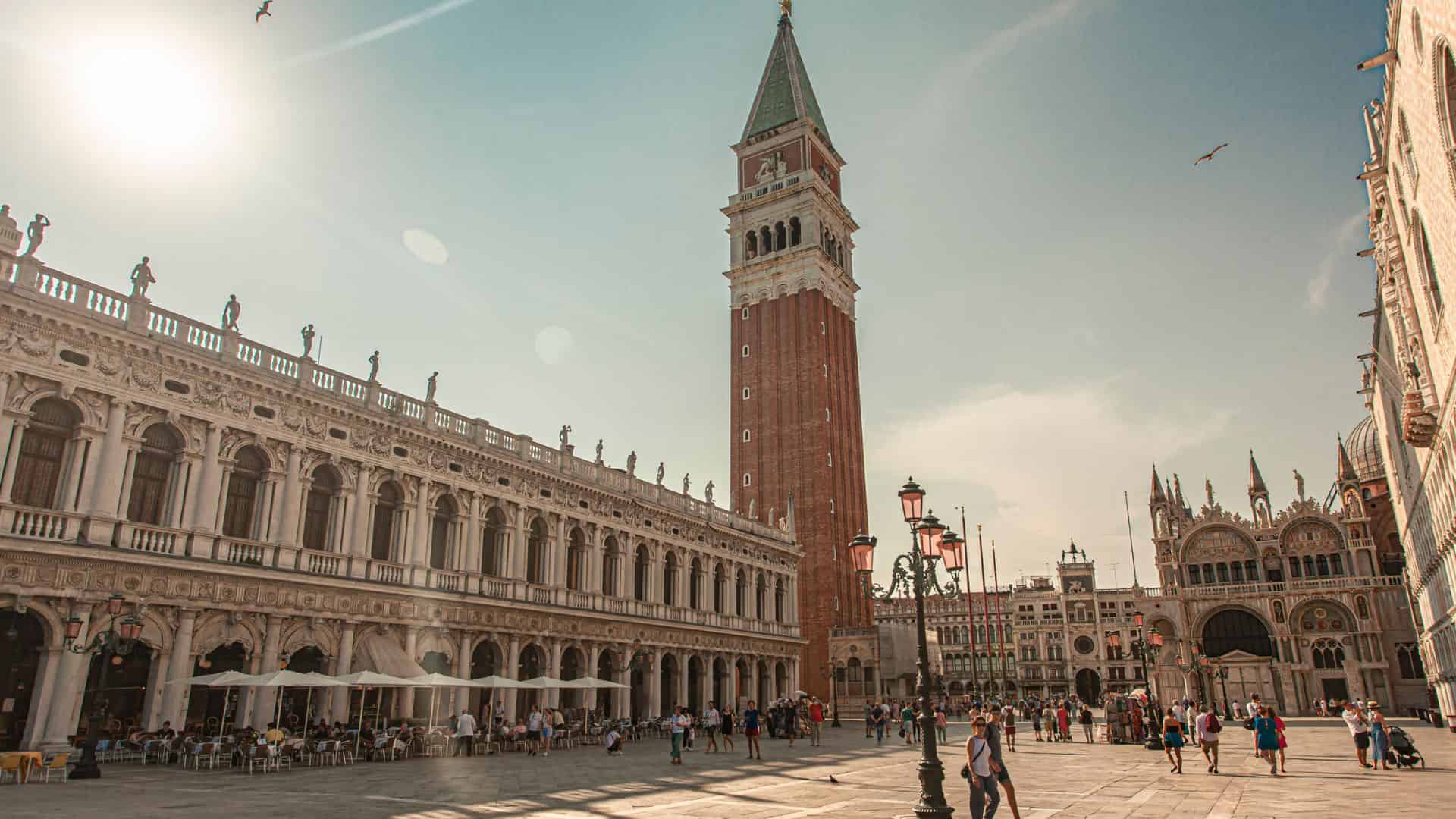 People walk in St. Mark's Square, Venice, with the Campanile and historic buildings under a sunny sky.