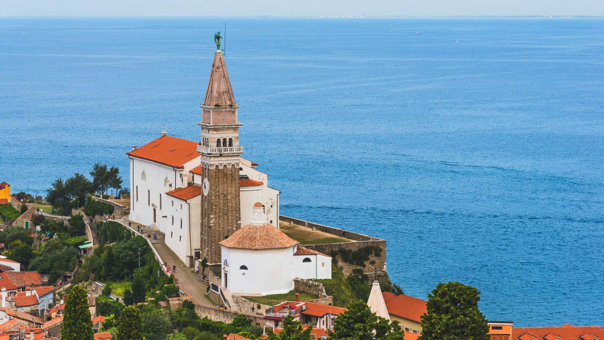 A coastal church with a tall bell tower overlooks the blue sea, surrounded by red-roofed buildings.
