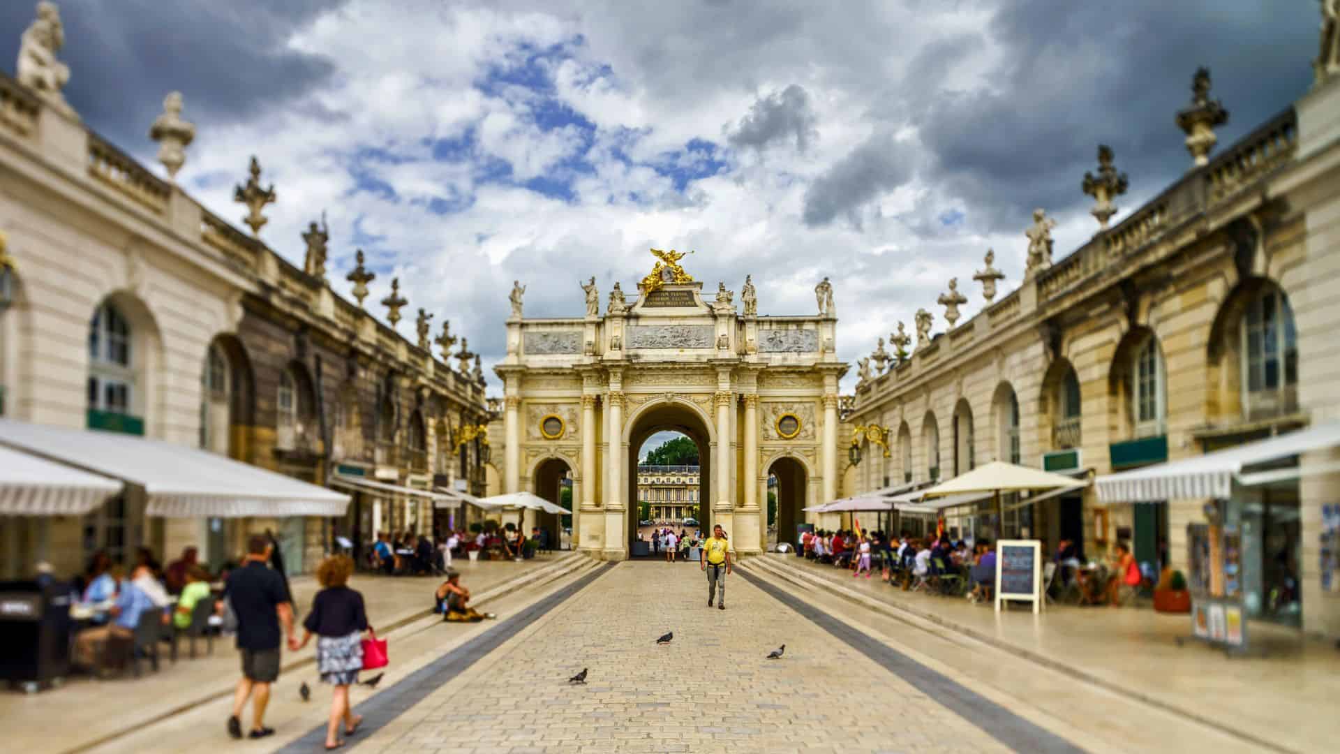 People walk and sit at outdoor cafés near an ornate archway under a cloudy sky in a European plaza.