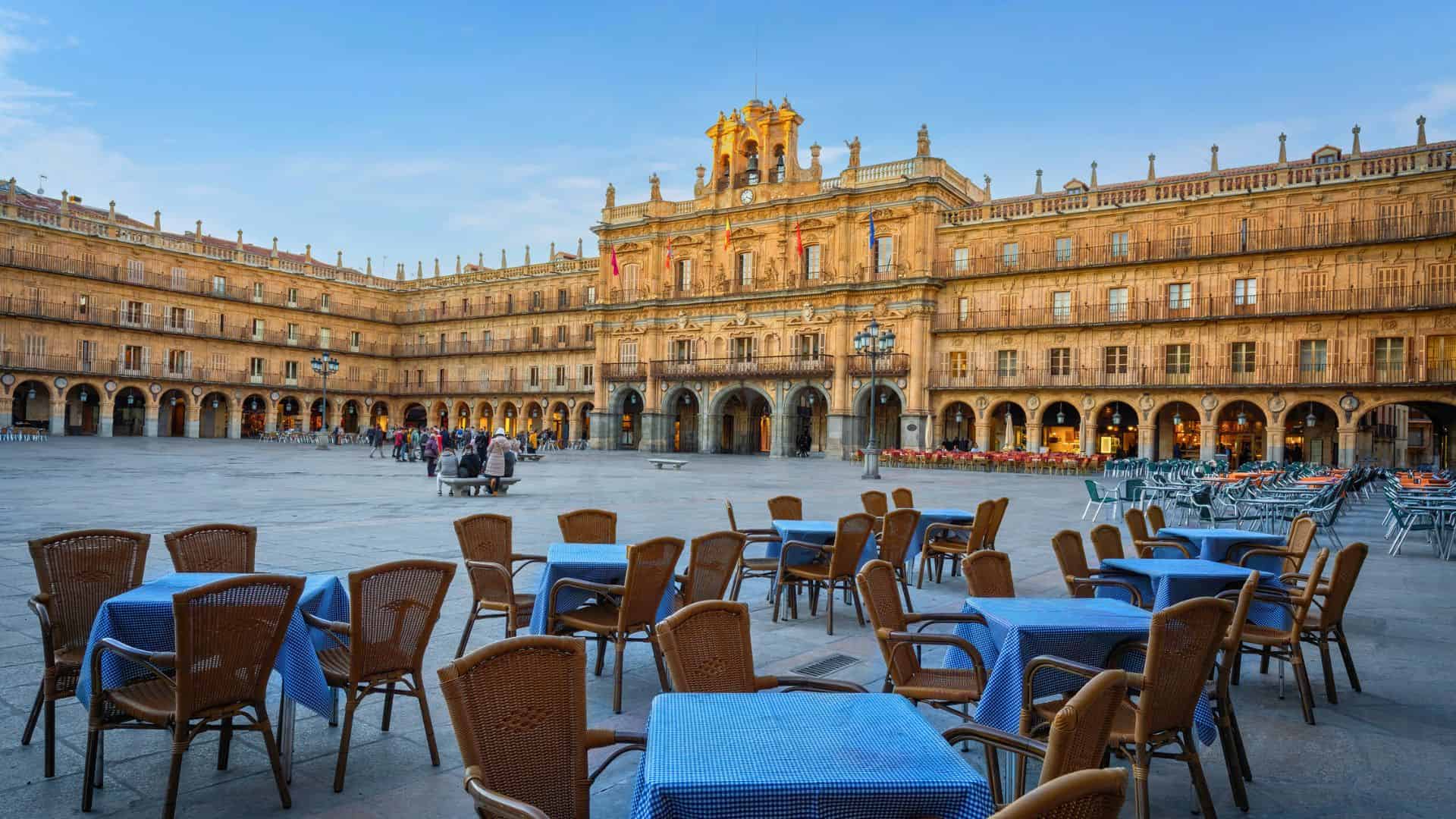 Outdoor café tables with blue cloths in a large, historic square surrounded by ornate buildings.
