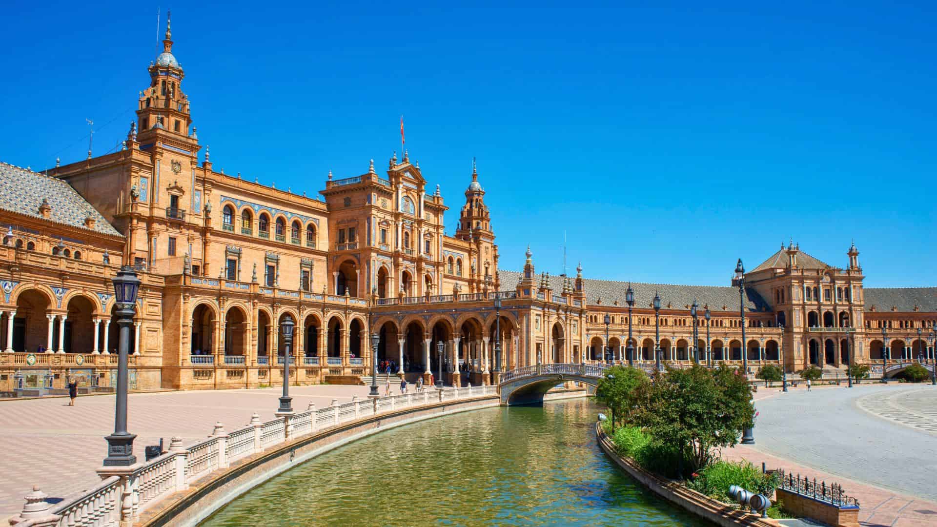 A grand, historic building with arches and towers beside a curved canal under a clear blue sky.